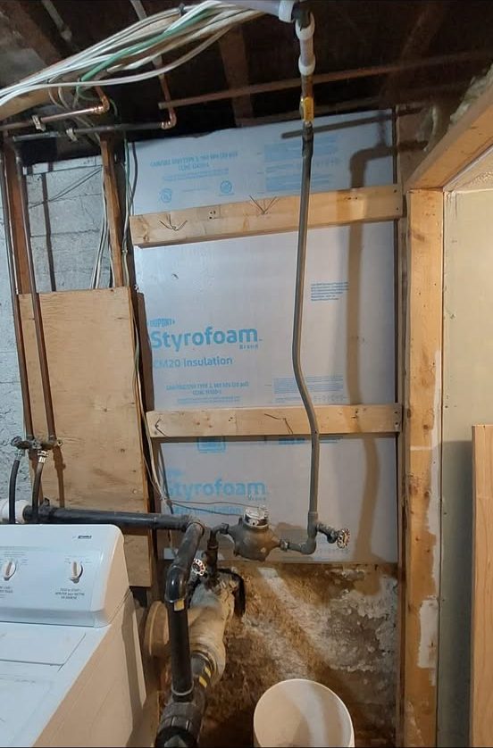 Old corroded water service entry and meter in a basement, with bare pipe and no backer board